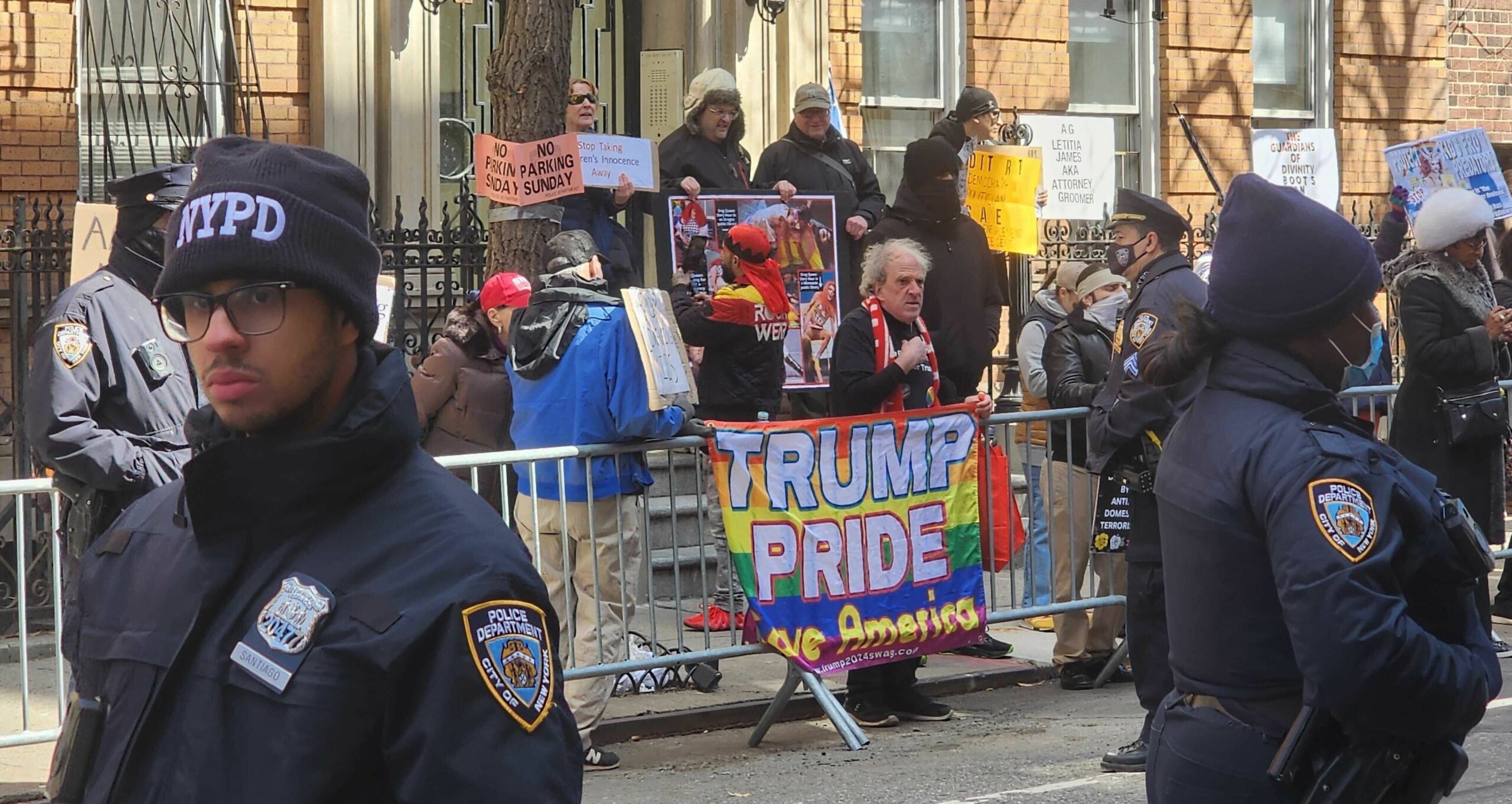 Une photo des manifestants contre la Drag Story Hour debout sur un trottoir, cernés par des clôtures métalliques mobiles placées le long du trottoir dans la rue. Une personne porte une épingle 